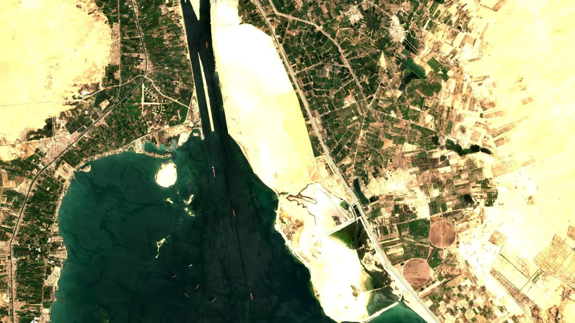 Sentinel-2 image of the Suez Canal entering the Great Bitter Lake in Egypt, with a few small ship shapes along the dark channel and green farmland and pale desert on either side.