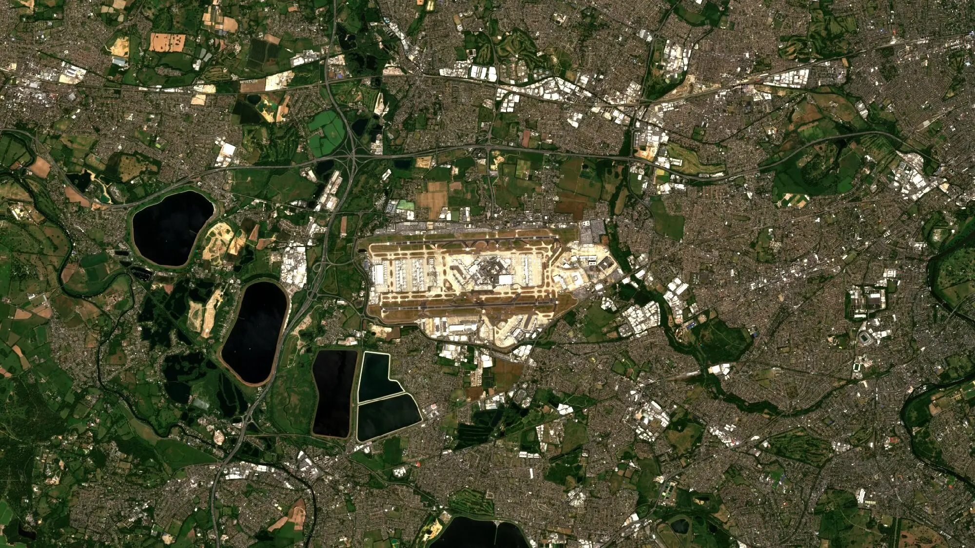 Sentinel-2 image of Heathrow Airport and west London, with the pale runways and terminals at the centre, dark reservoirs to the west and motorways crossing green fields and dense suburbs.
