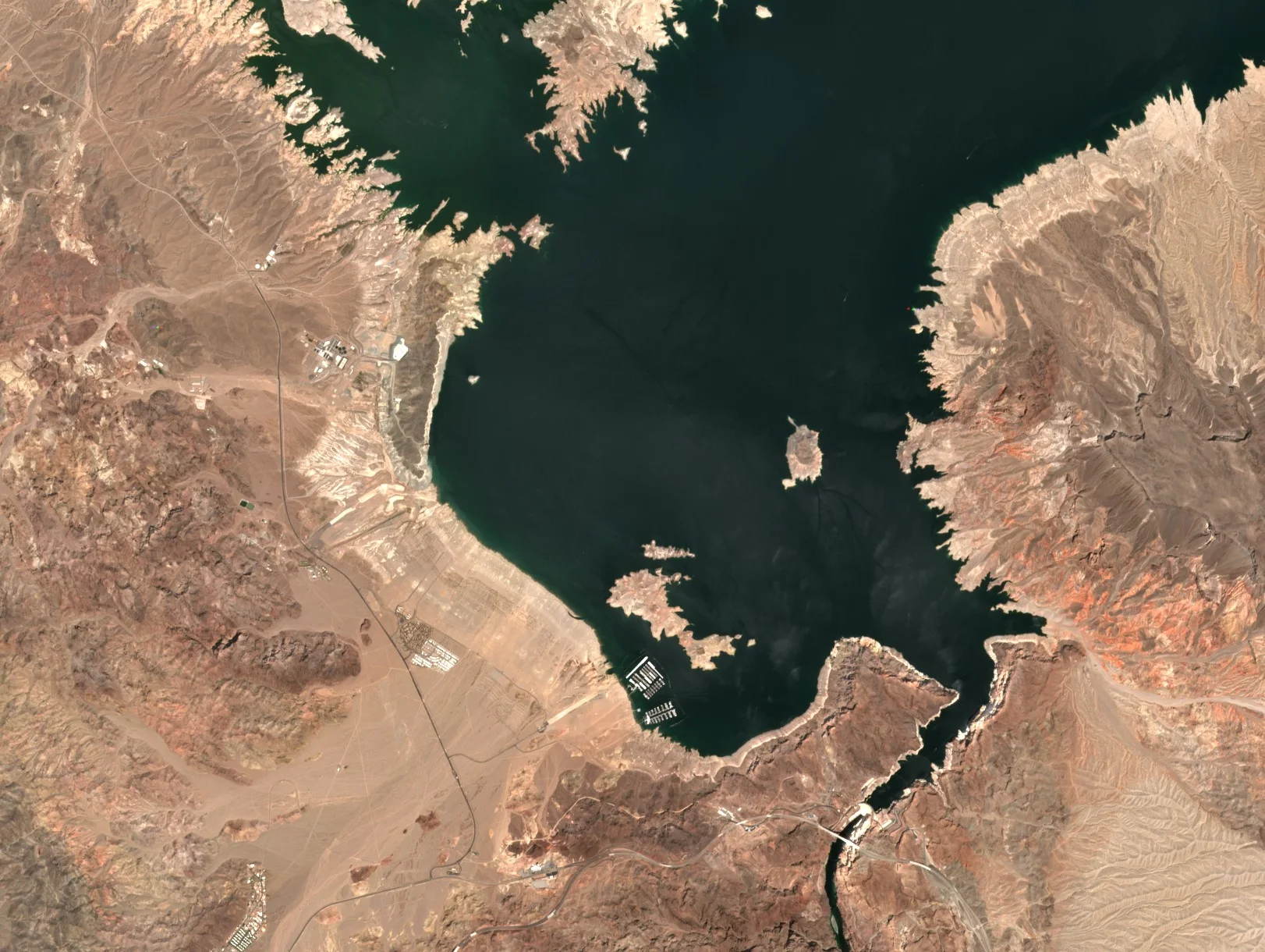 A large dark lake among red and tan desert mountains, ringed by a wide pale band of exposed shore, with a dam and a narrow canyon at the bottom of the image.