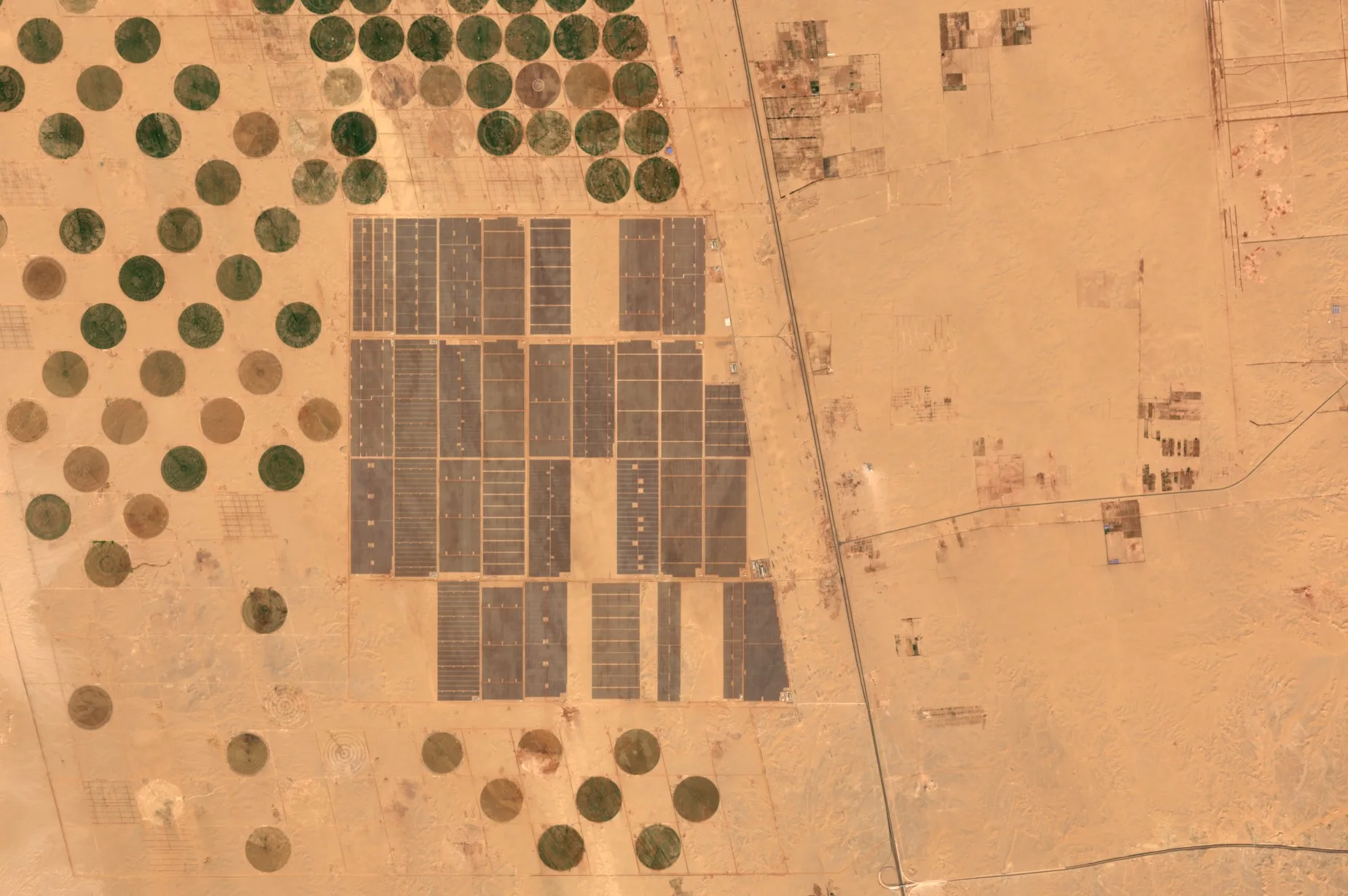 Pale desert with a large grid of dark rectangles at the centre, each a block of solar panels, and green and brown circular fields to the west and south.