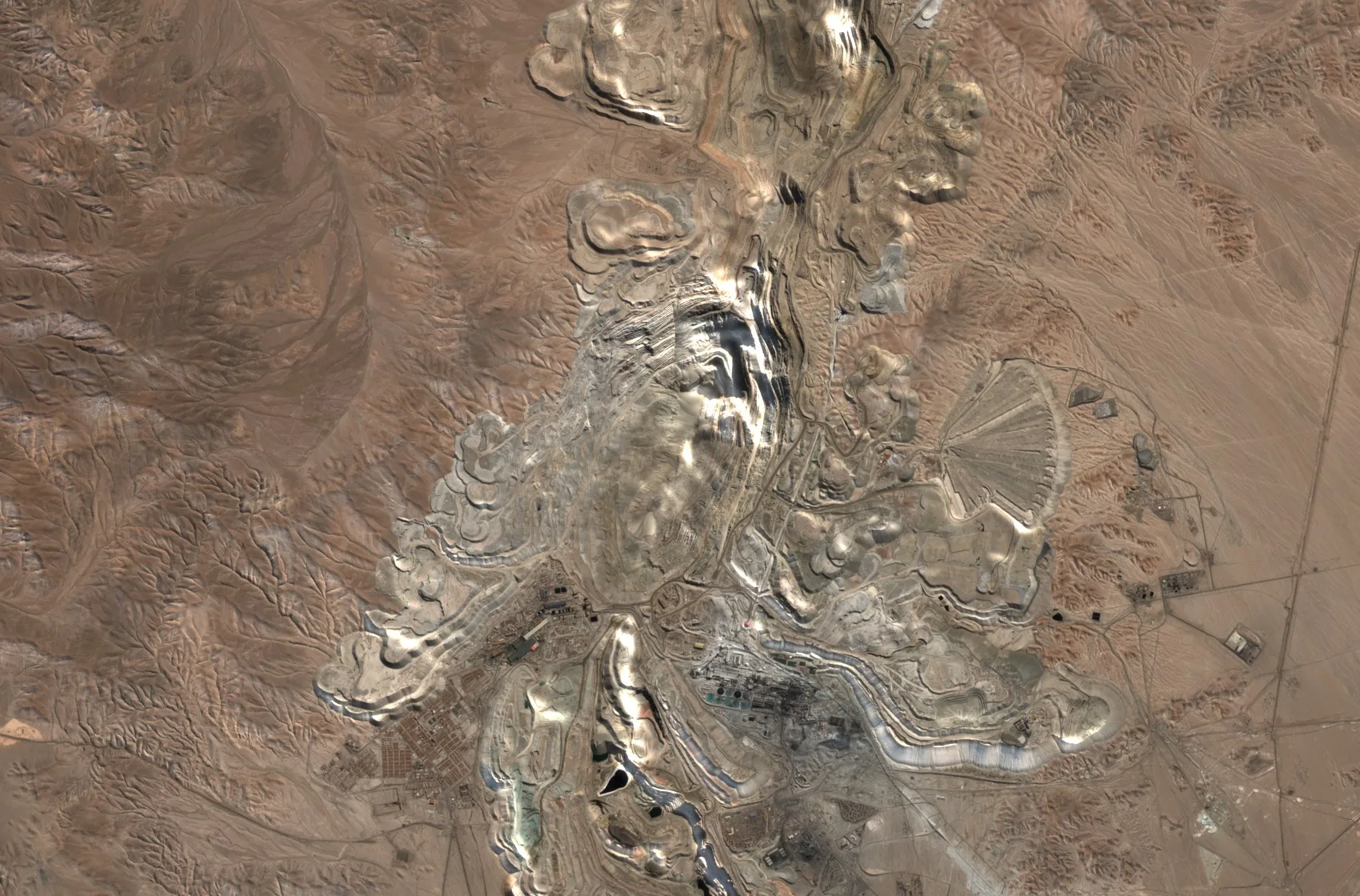 A large copper mine in brown desert. A huge pale open pit with terraced benches lies at the centre, ringed by grey waste rock heaps with stepped edges, with buildings and a processing plant to the south and bare desert hills around.