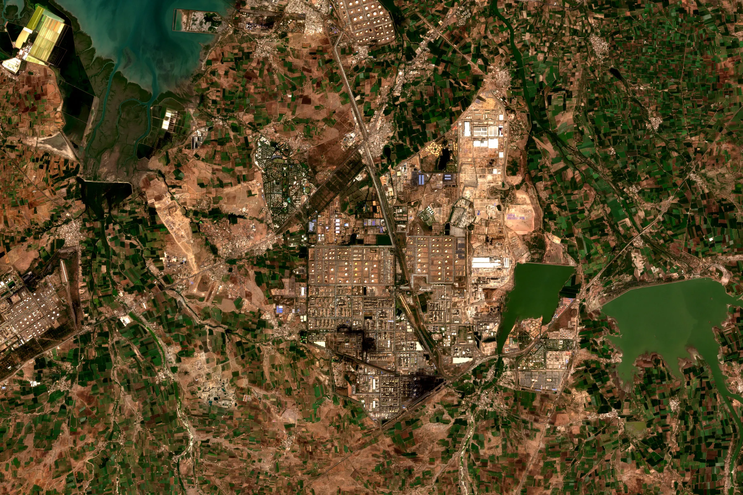 Satellite view of the Jamnagar refinery and petrochemical complex in Gujarat, India, with grids of storage tanks and process units among farmland, a green reservoir to the east and a turquoise bay to the north-west